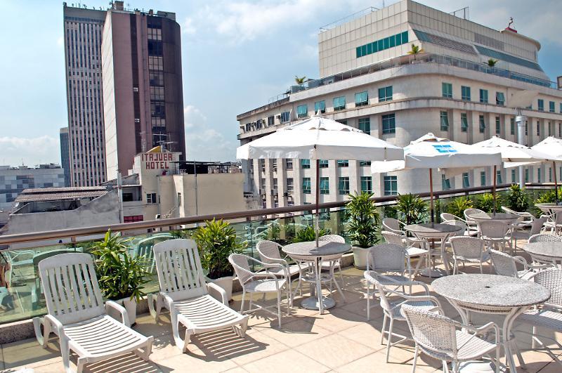 호텔 아틀란티코 비즈니스 센트로 리오데자네이루 외부 사진 The photo shows a rooftop area with several white lounge chairs and circular tables. There are umbrellas providing shade over some of the seating. In the background, there are tall buildings, including a modern skyscraper with a distinct architectura